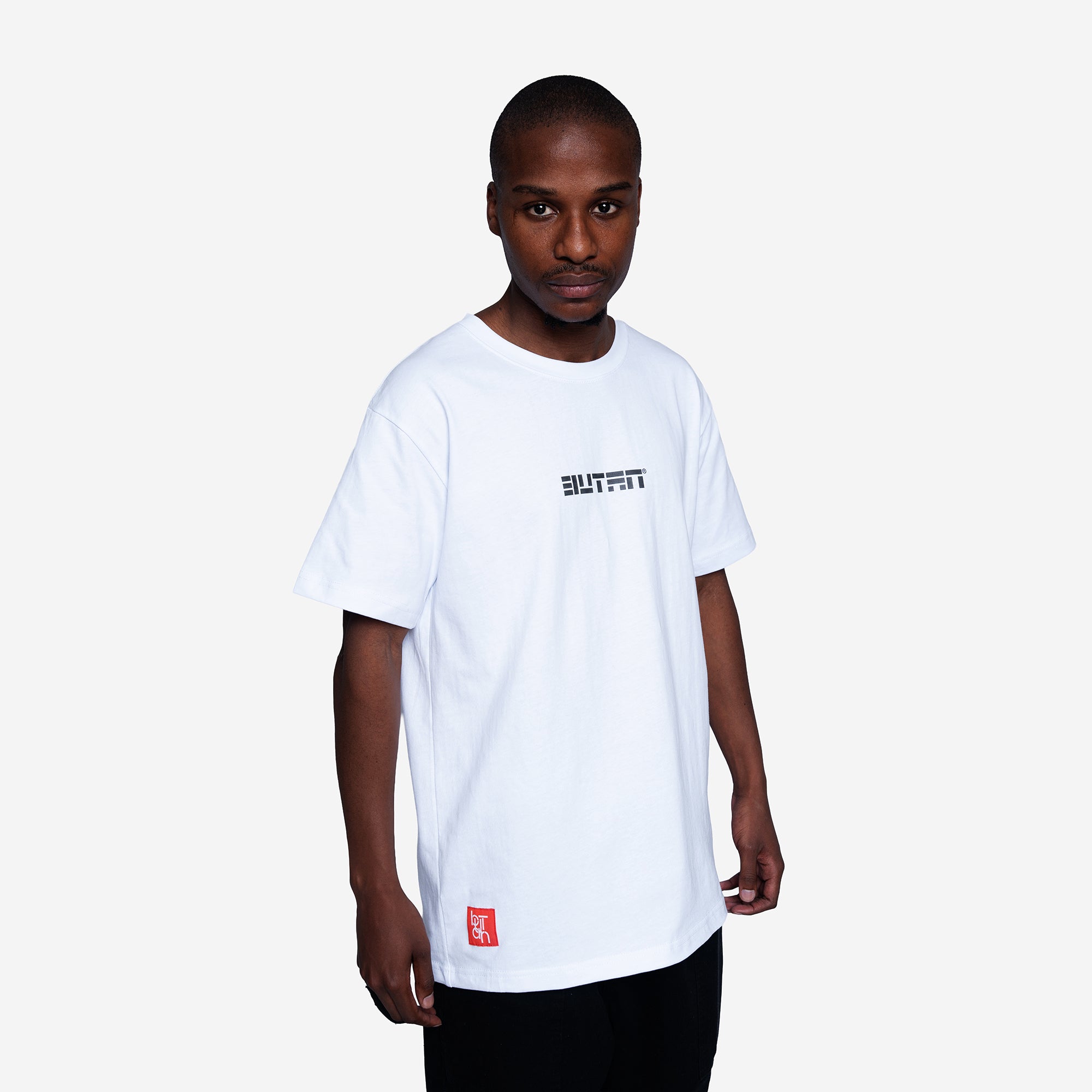 A man wearing a white short-sleeve crewneck t-shirt with a black 'BUTAN' logo on the chest and a small red square graphic near the bottom hem, set against a plain light gray background.