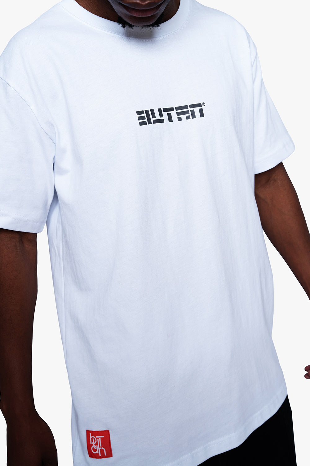 A white short-sleeve t-shirt with a black 'BUTAN' logo printed on the chest and a small red square patch near the bottom hem, worn by a model.