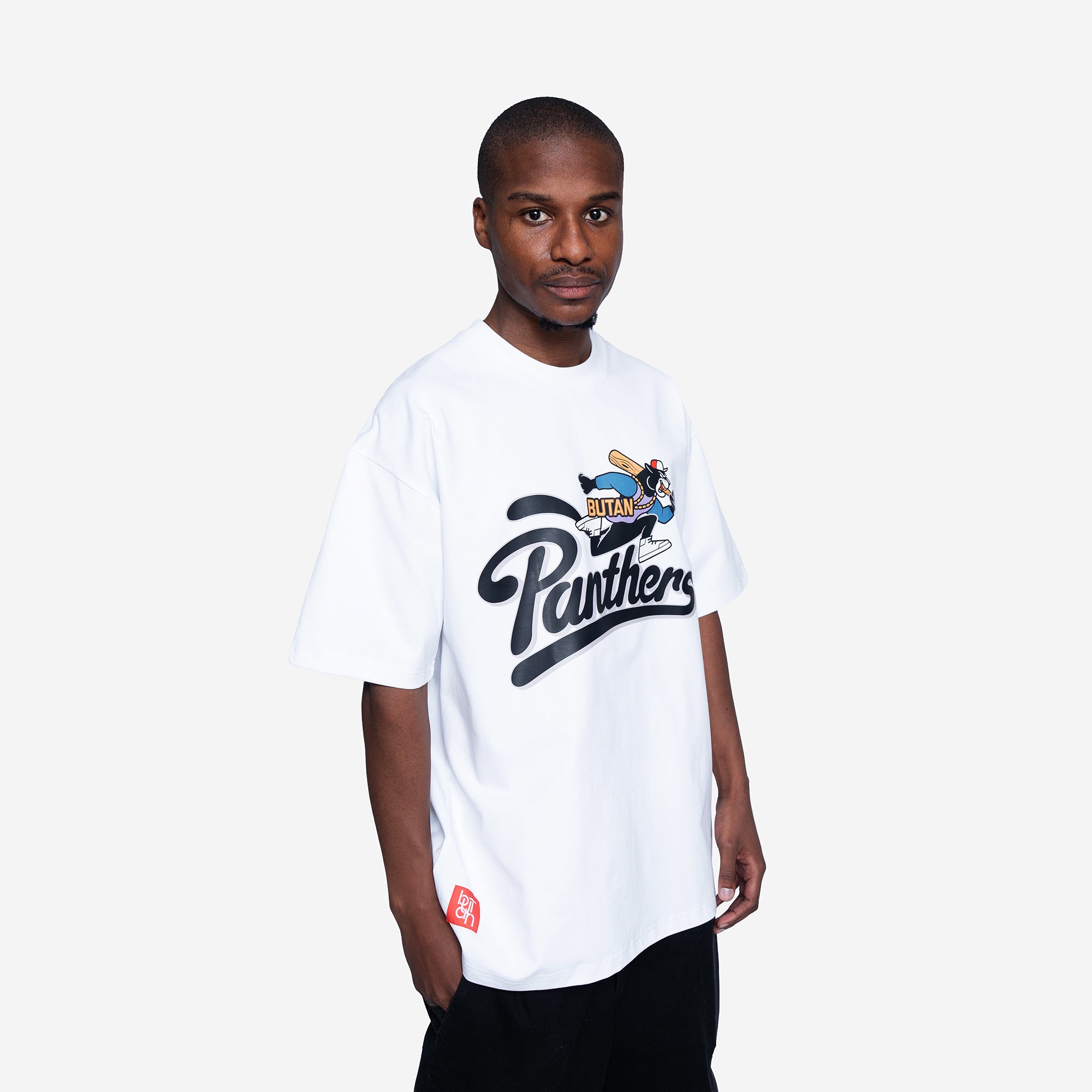 A man wearing a white oversized t-shirt with a black Panthers graphic and a bee logo on the chest, paired with black pants, against a light gray background.