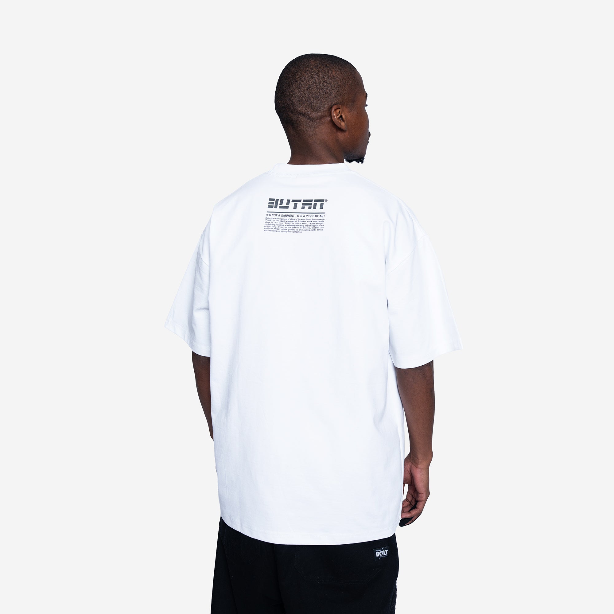 A man viewed from behind wearing a white oversized t-shirt with a black graphic print on the upper back and black pants, set against a plain light gray background.