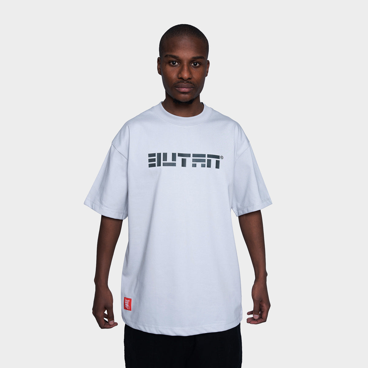 A man wearing a white short-sleeve t-shirt with a black 'BUTAN' graphic across the chest and a small red tag near the hem, set against a plain light gray background.