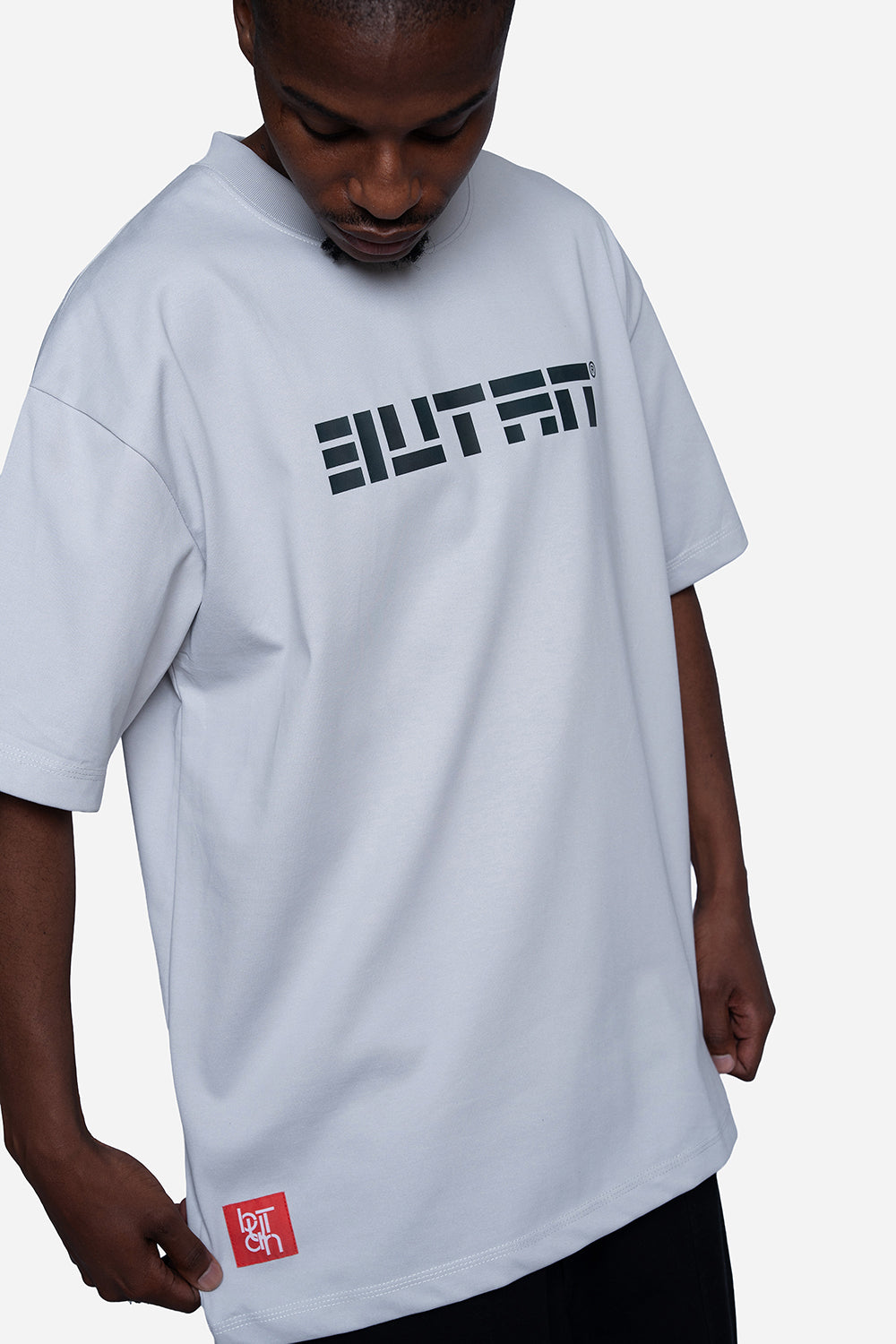 A man wearing an oversized white t-shirt with a black stylized 'BUTAN' logo across the chest and a small red square logo near the bottom hem, set against a plain light gray background.