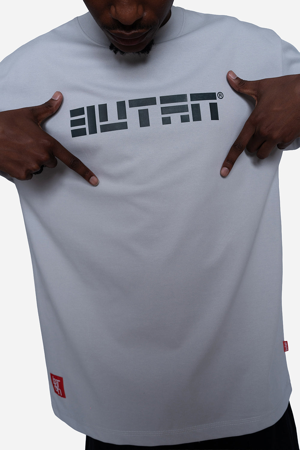 A close-up of a person wearing a light gray t-shirt with a bold black 'BUTAN' logo printed across the chest, pointing at the logo with both hands.