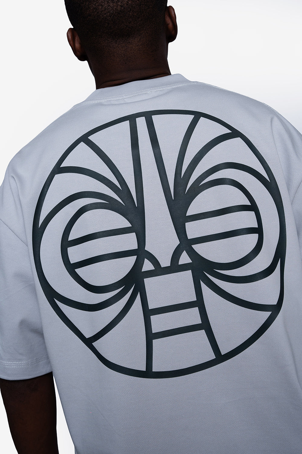 A man viewed from behind wearing a white t-shirt with a large black tribal face graphic printed on the back.