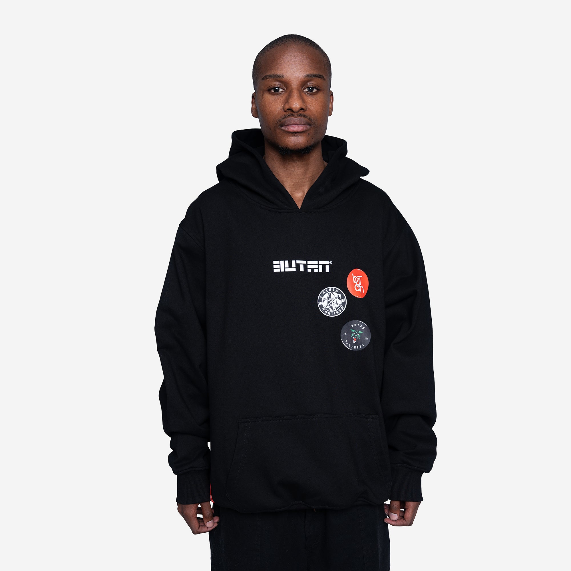 A model wearing a black pullover hoodie with 'BUTAN' printed in white across the chest, alongside three circular graphic patches, against a light gray background.