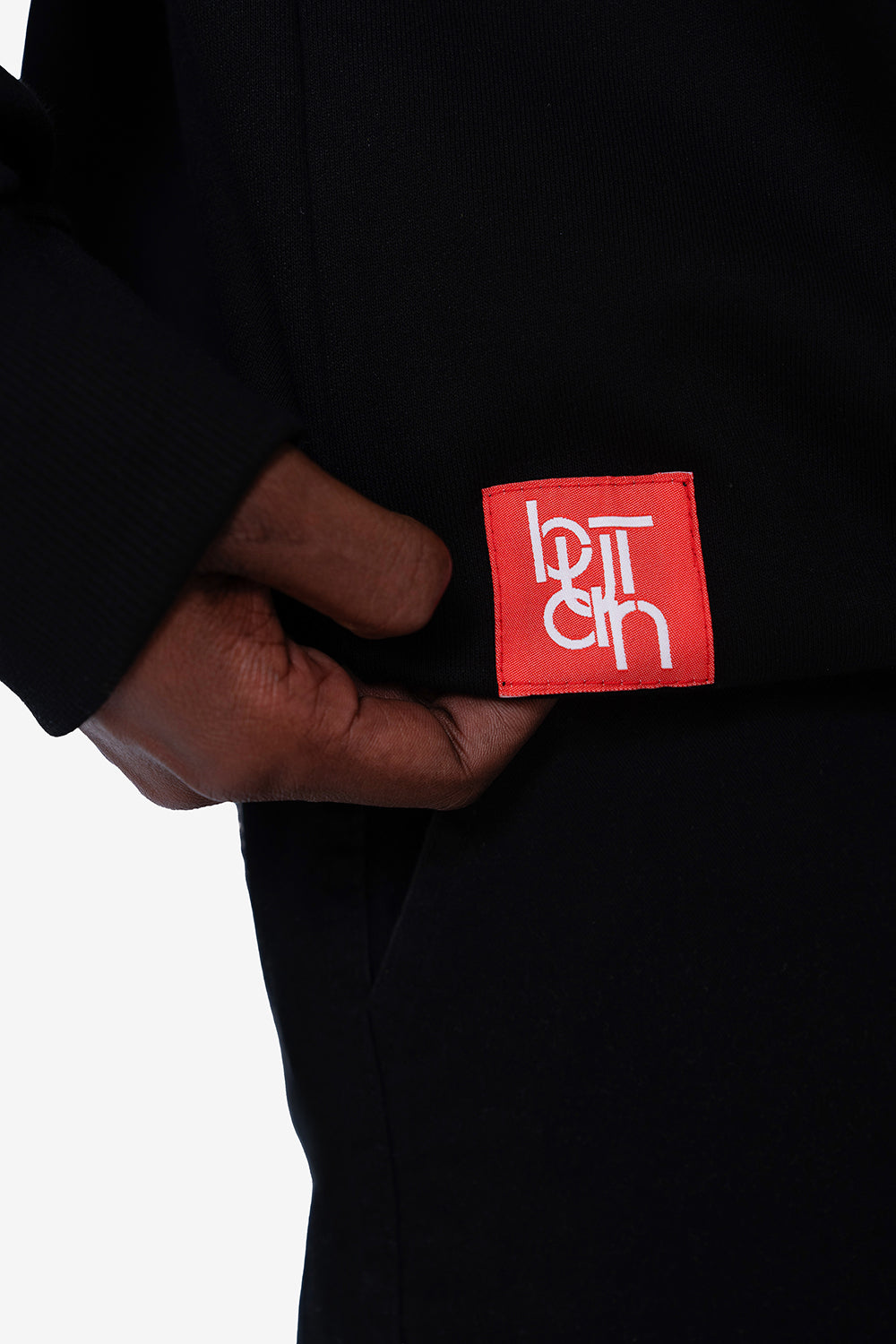 A close-up of a person wearing a black jacket or sweatshirt, pulling a red square patch from the pocket. The patch features the brand name 'BUTAN' in stylized white lettering.