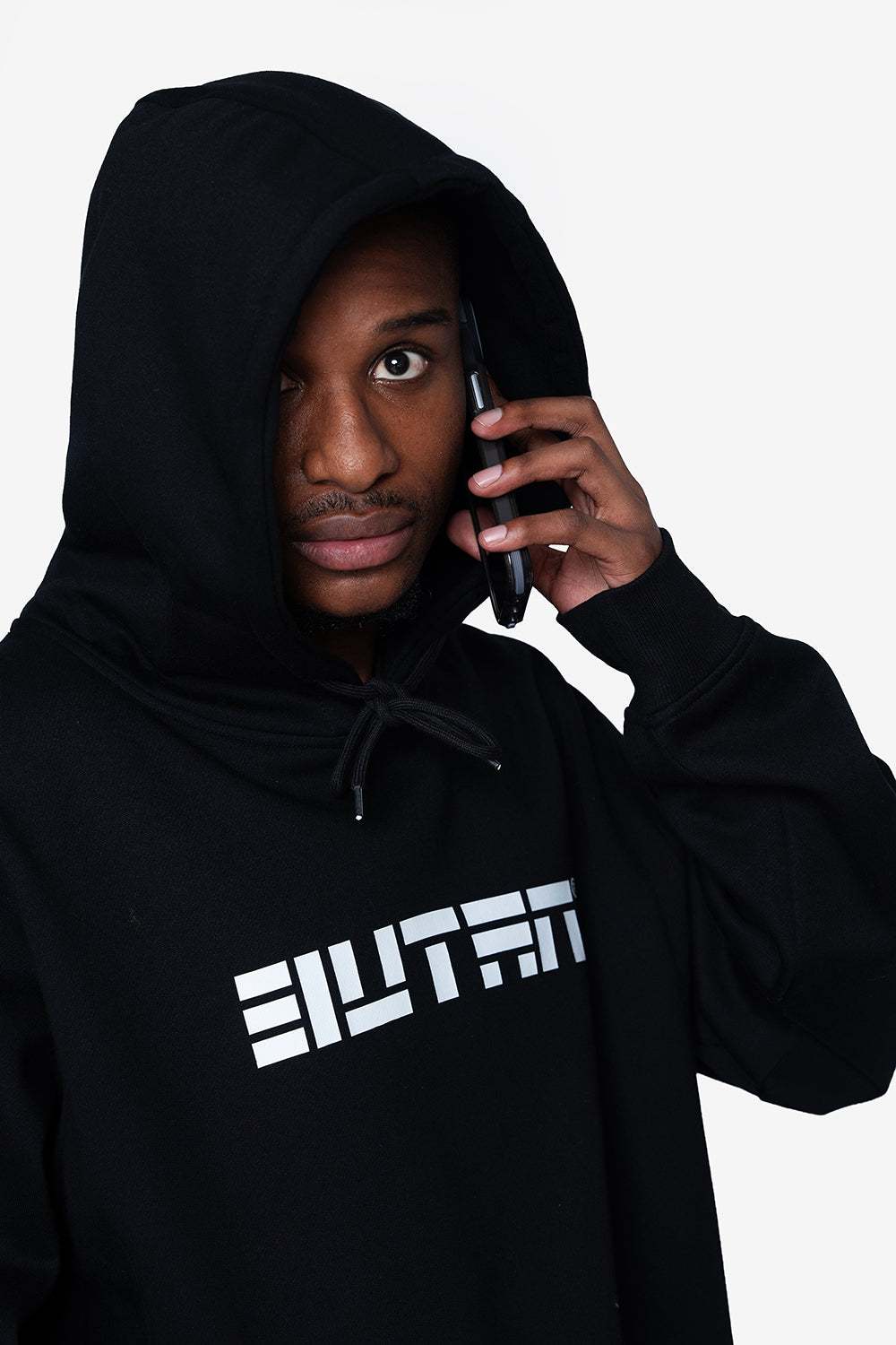 A man wearing a black pullover hoodie with a white stylized 'BUTAN' logo on the chest, holding a black smartphone to his ear against a light gray background.