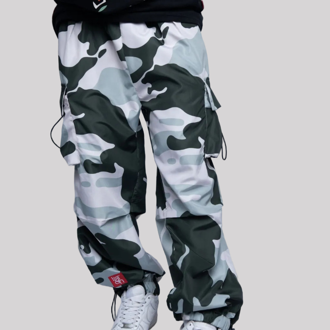 Person wearing camouflage pants and a black hoodie with visible branding on a light gray background