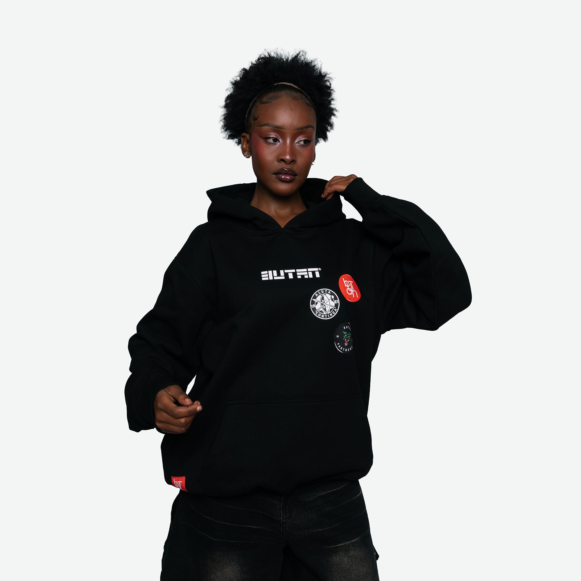 A model wearing a black pullover hoodie with 'BUTAN' printed in white on the chest, alongside several circular and rectangular patches, set against a plain light gray background.