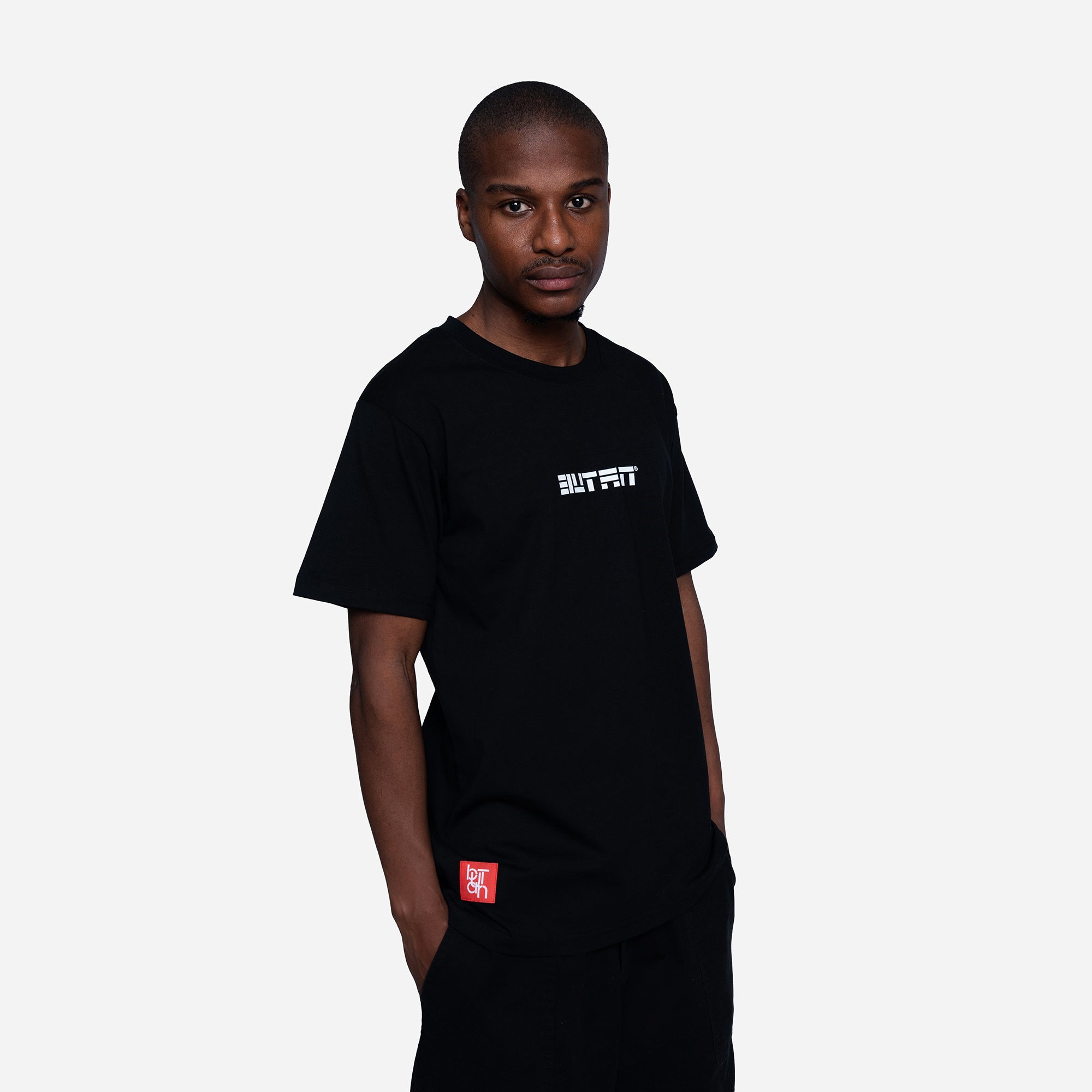 A man wearing a black short-sleeve t-shirt with white text on the chest and a red square graphic on the pocket, photographed against a light gray background.