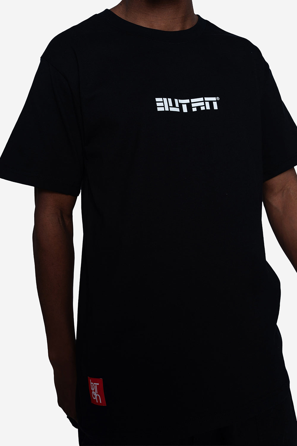 A person wearing a black short-sleeve t-shirt with a white stylized 'BUTAN' logo printed on the chest and a small red square patch near the lower left hem, shown against a plain light background.