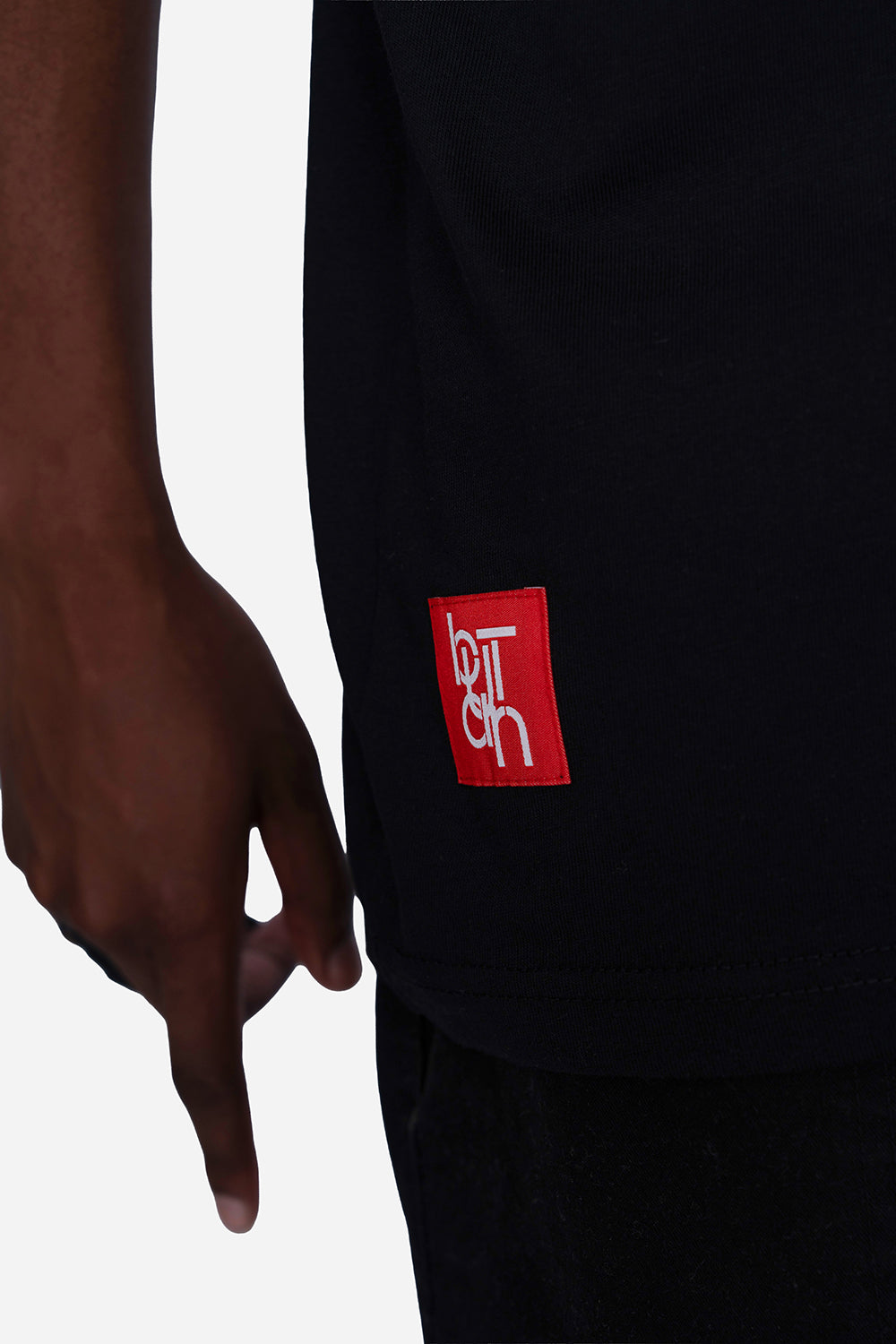 Close-up of a person wearing a black T-shirt with a red square patch featuring stylized white lettering on the lower side, against a light gray background.
