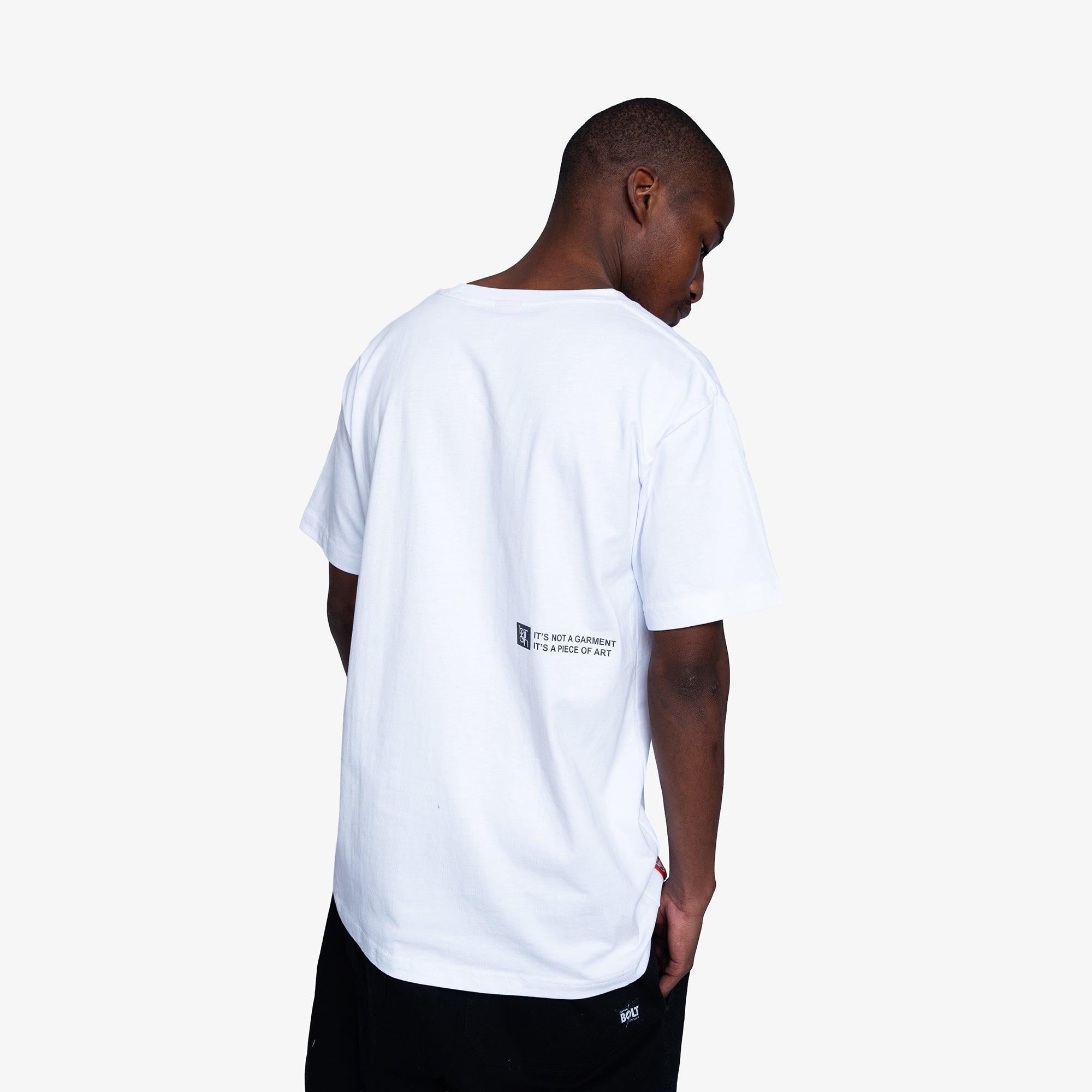 A man viewed from behind wearing a white oversized t-shirt with a small black graphic on the back and black shorts, set against a plain light gray background.