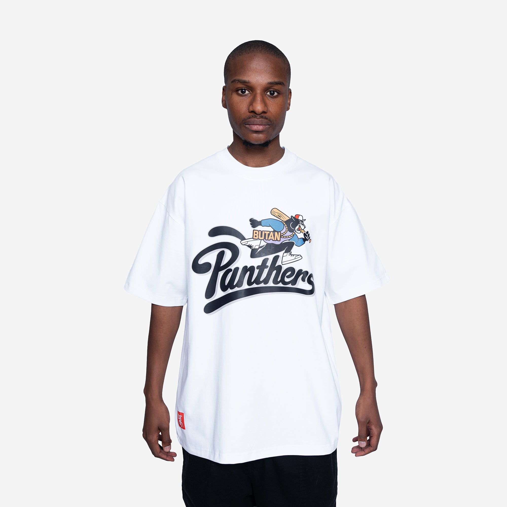 A man wearing a white oversized t-shirt with a black and blue 'Panthers' graphic on the front, paired with black pants, against a light gray background.