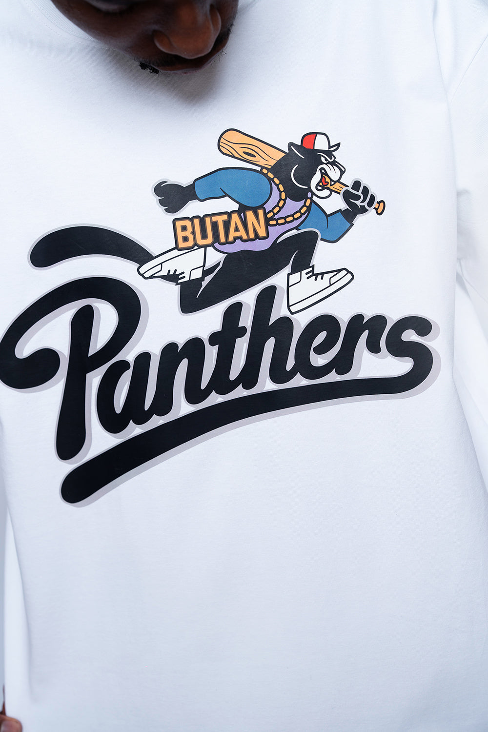 A close-up of a white long-sleeve shirt featuring a large graphic print. The design displays a cartoon black panther wearing a blue cap and gold chain, holding a bat, with the word 'BUTAN' in yellow.