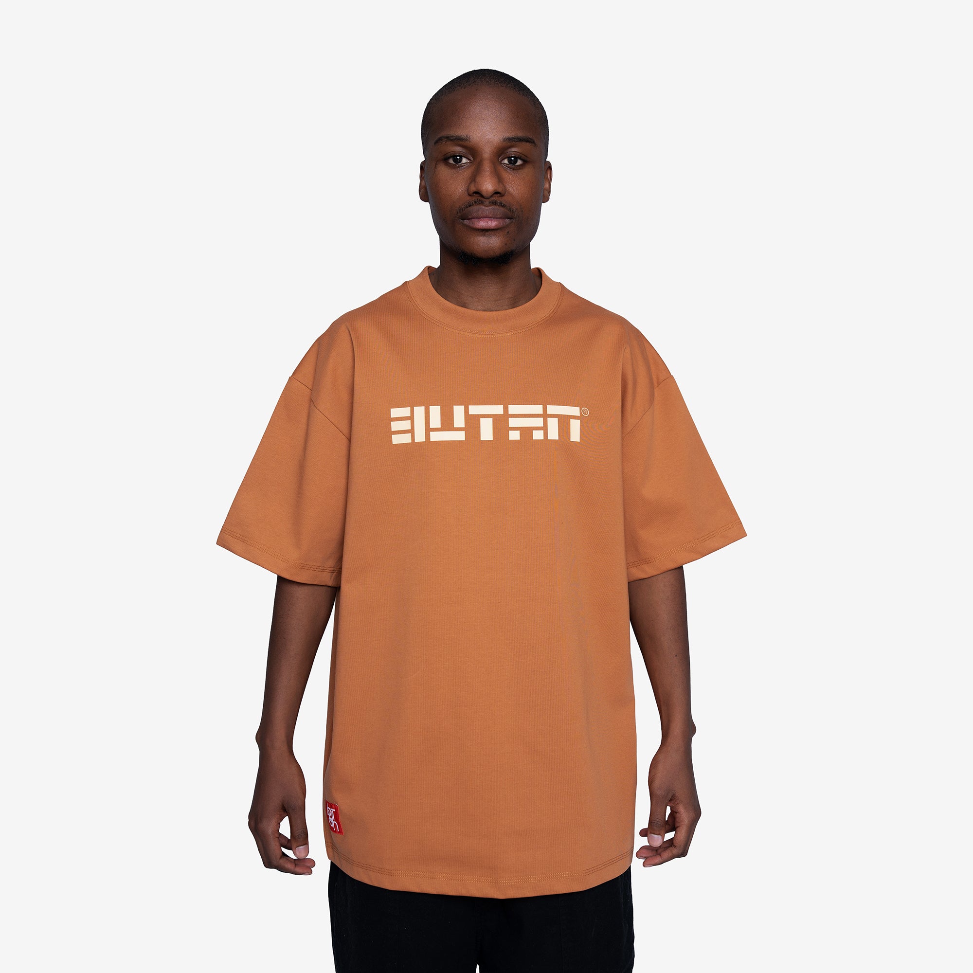 A man wearing an oversized orange crewneck t-shirt with the word 'BUTAN' printed in white across the chest, photographed against a light gray background.