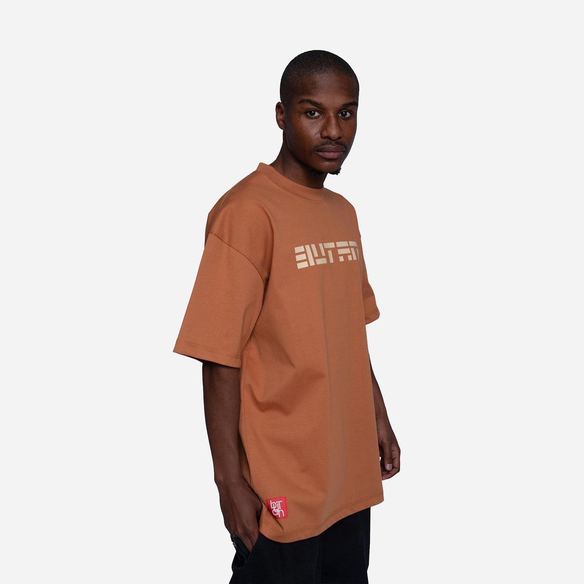 A man wearing a rust-orange oversized t-shirt with a beige graphic print on the chest and a small red tag near the hem, photographed against a light gray background.