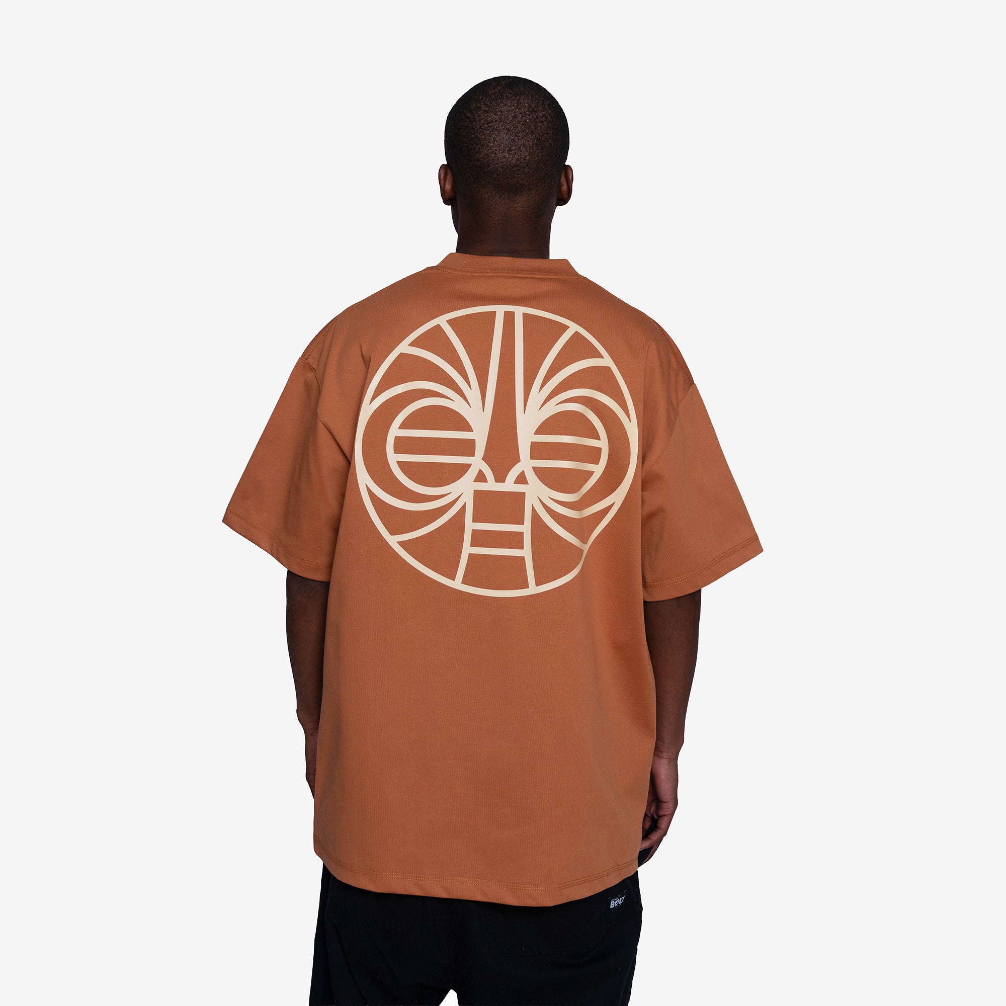 A man viewed from behind wearing a rust-orange t-shirt with a large cream-colored circular graphic print on the back, paired with black pants, against a light gray studio background.