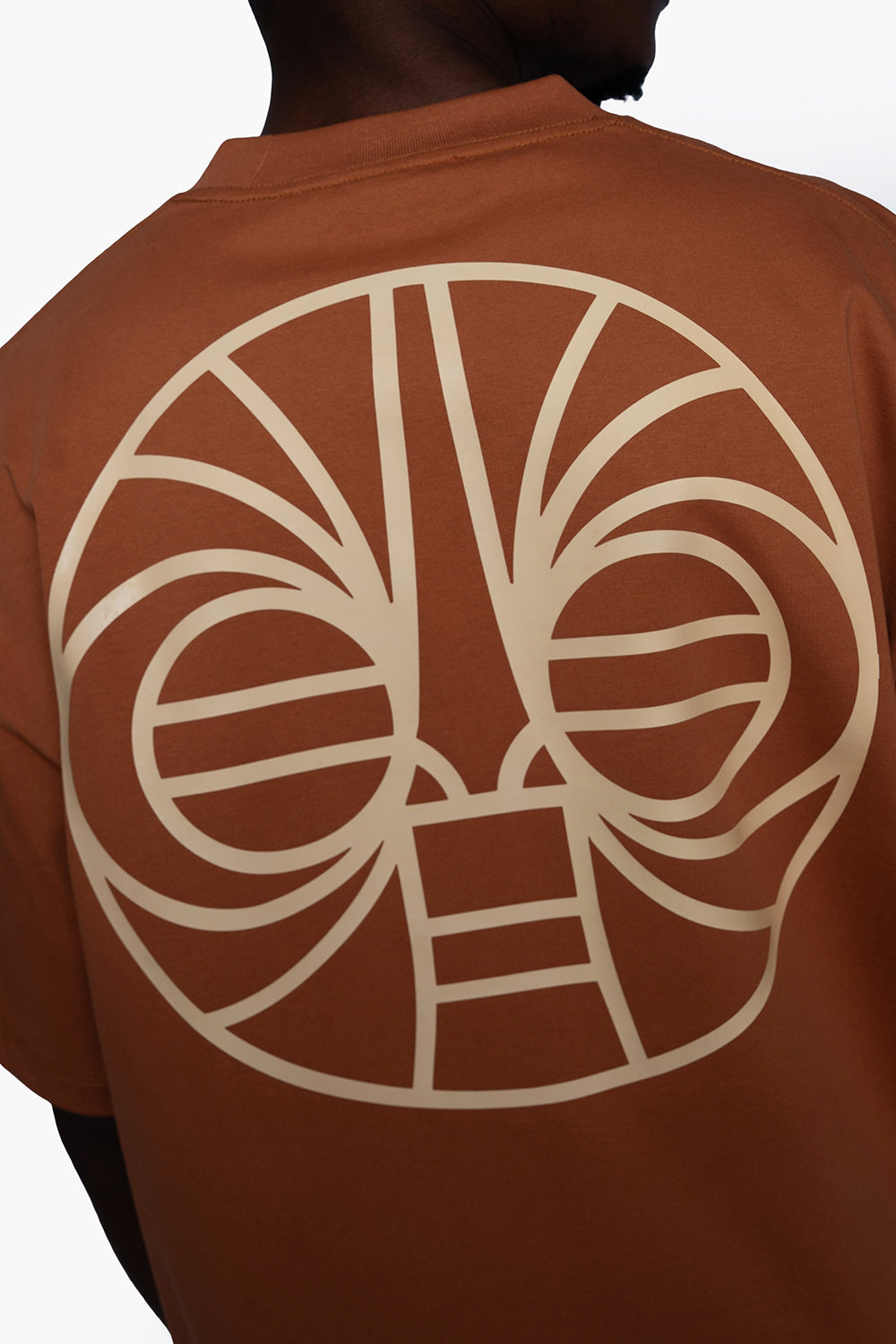 A close-up of a person's back wearing a rust-brown t-shirt with a large cream-colored tribal face graphic printed on the back.