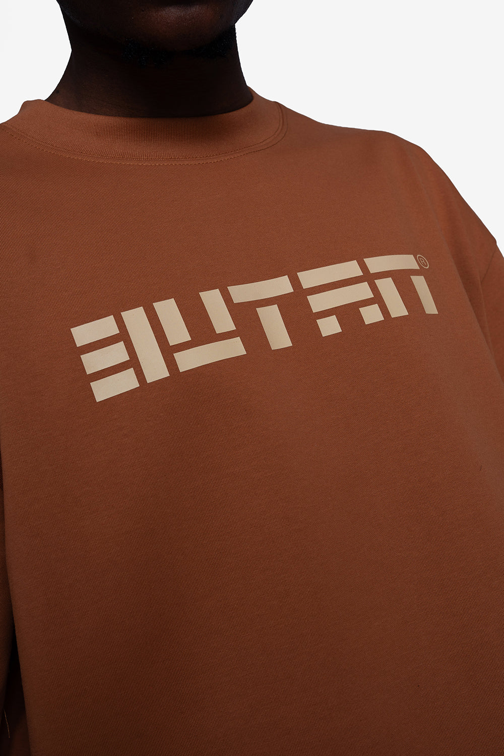 A close-up of a person wearing a brown crewneck sweatshirt with a large beige stylized graphic logo across the chest.