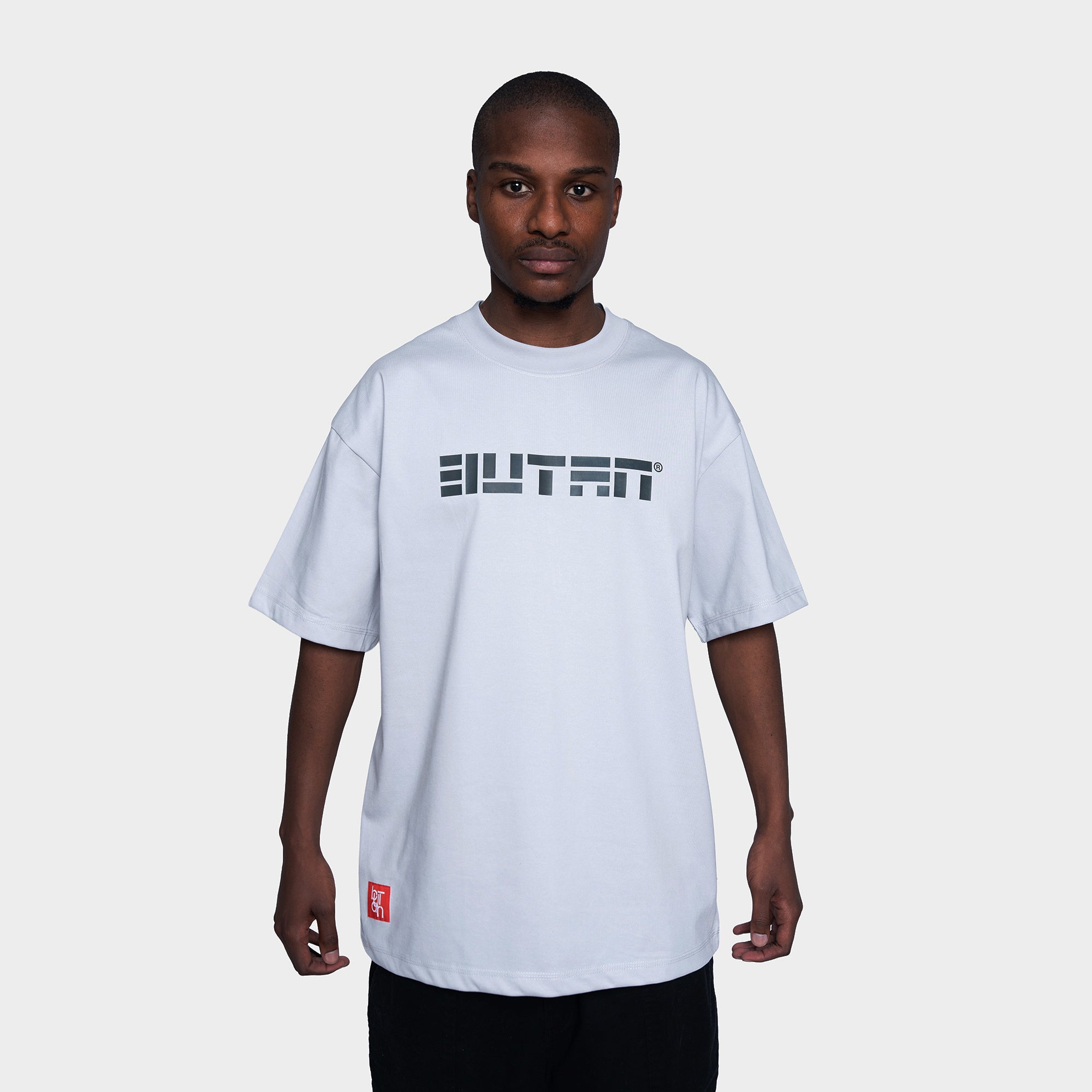 A man wearing a white short-sleeve t-shirt with a black 'BUTAN' graphic across the chest and a small red tag near the hem, set against a plain light gray background.