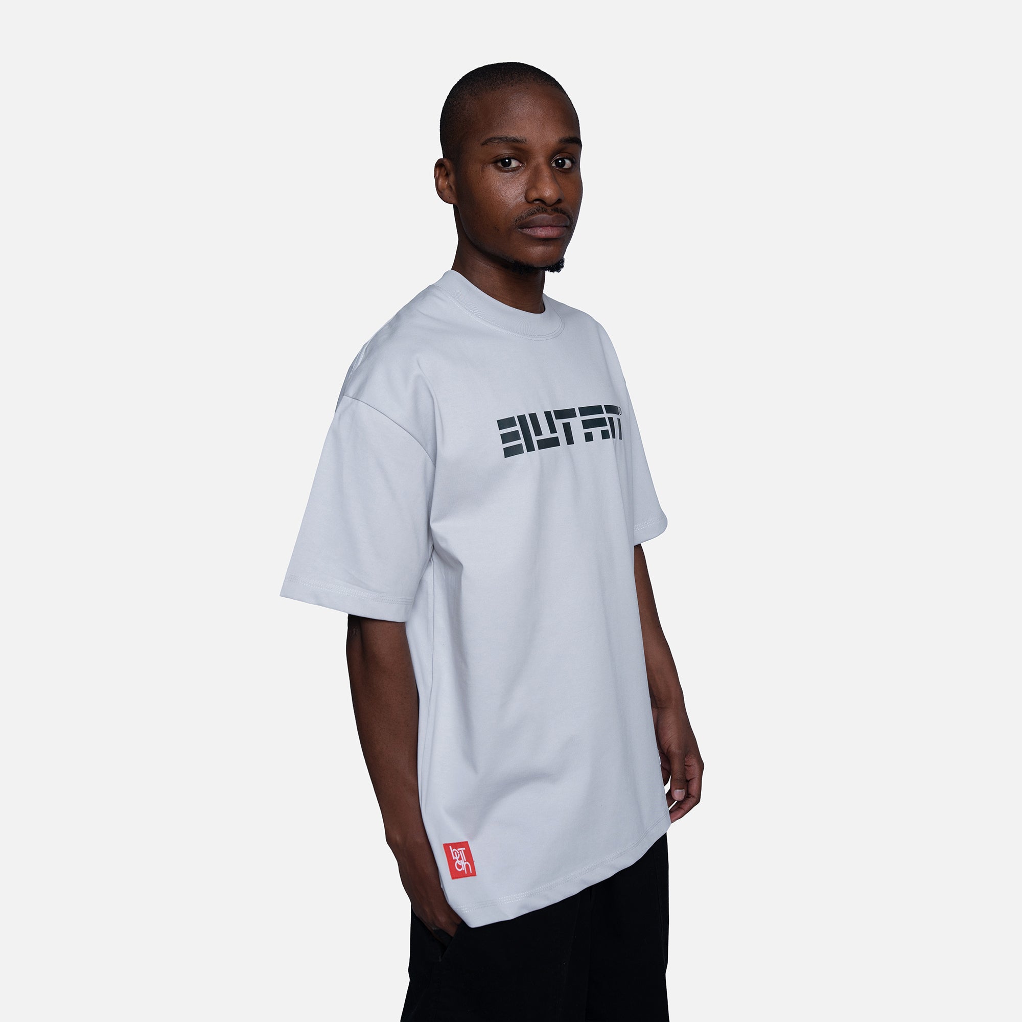 A man wearing a light gray oversized t-shirt with a black 'BUTAN' graphic across the chest and a small red tag near the bottom hem, set against a plain light gray background.
