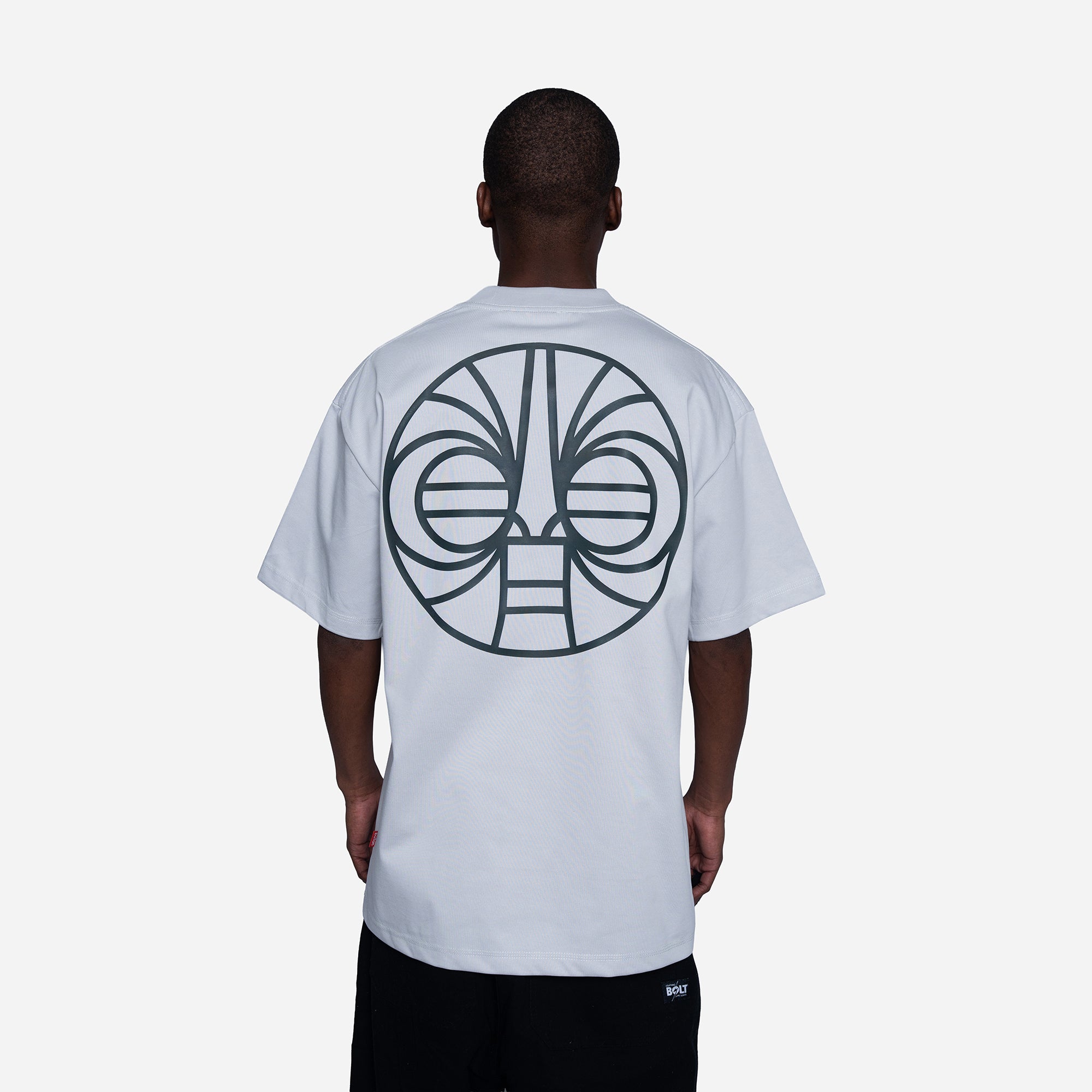 A man viewed from behind wearing a white t-shirt with a large black tribal face graphic printed on the back, paired with black pants, against a light gray studio background.