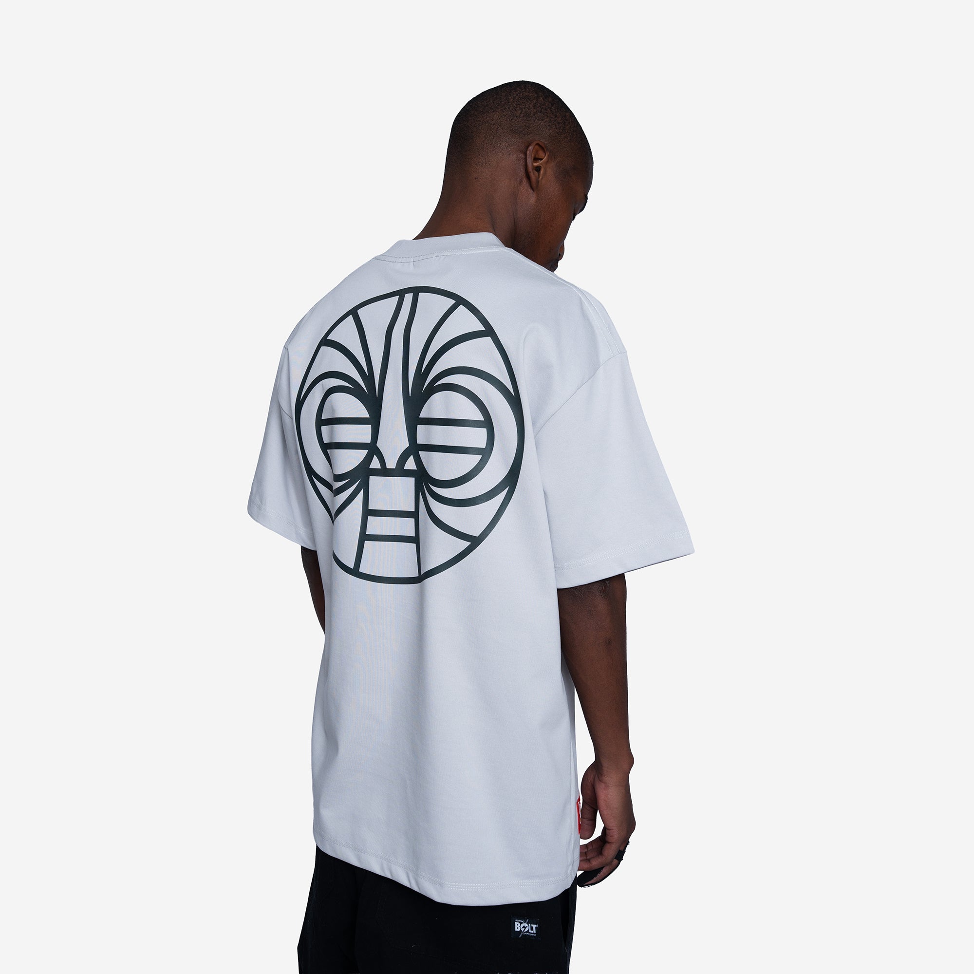 A man viewed from behind wearing a white t-shirt with a large black graphic print of a tribal face on the back, paired with black shorts against a light gray background.
