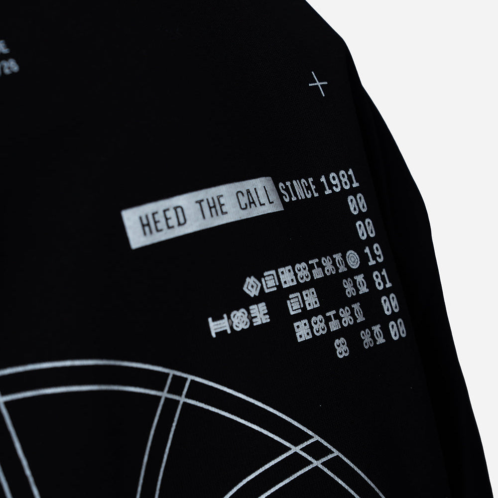 Close-up of a black garment with white printed text reading 'HEED THE CALL SINCE 1981' and white curved line graphics on a light gray background.