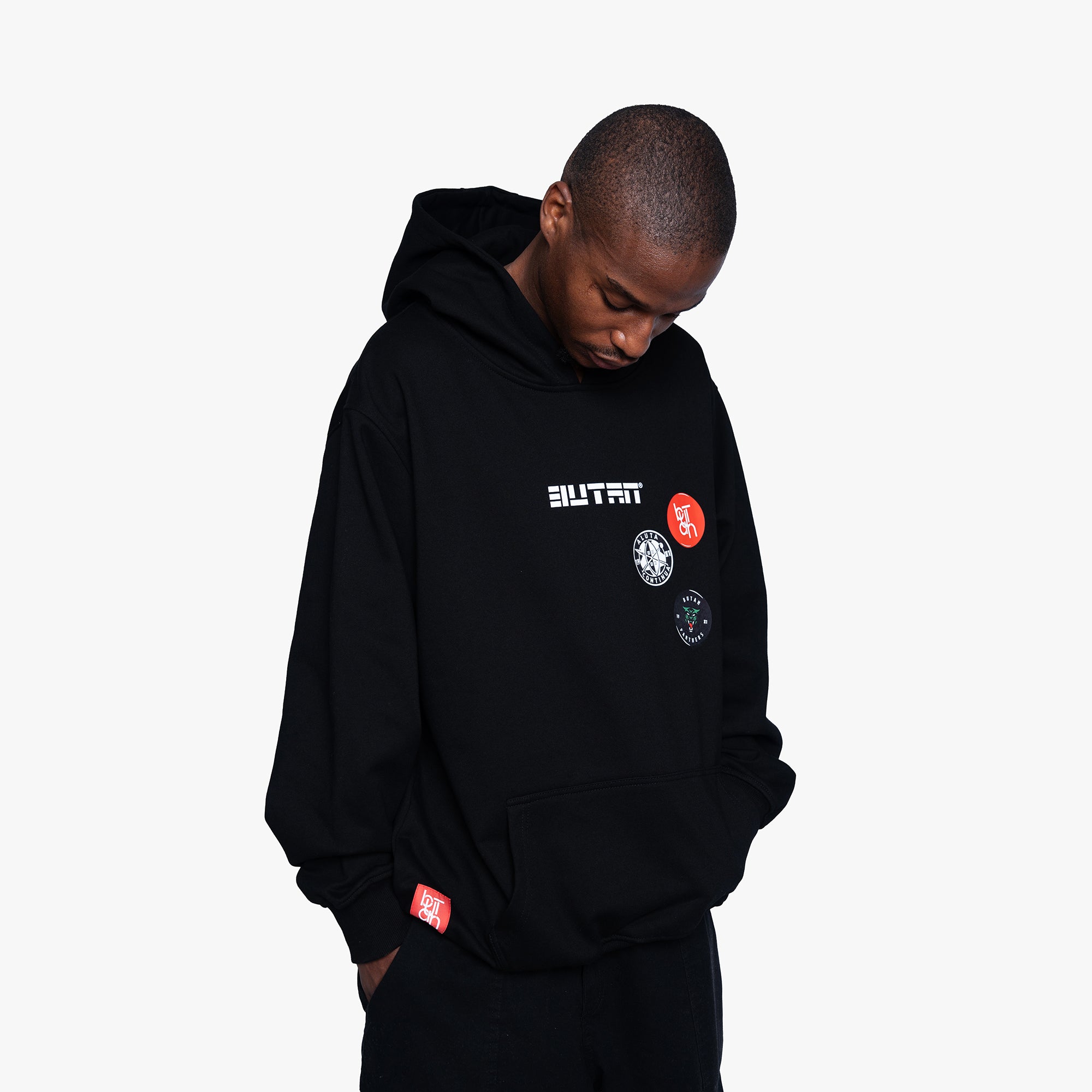 A man wearing a black pullover hoodie with 'BUTAN' printed in white on the chest, accompanied by several circular graphic patches, photographed against a light gray background.