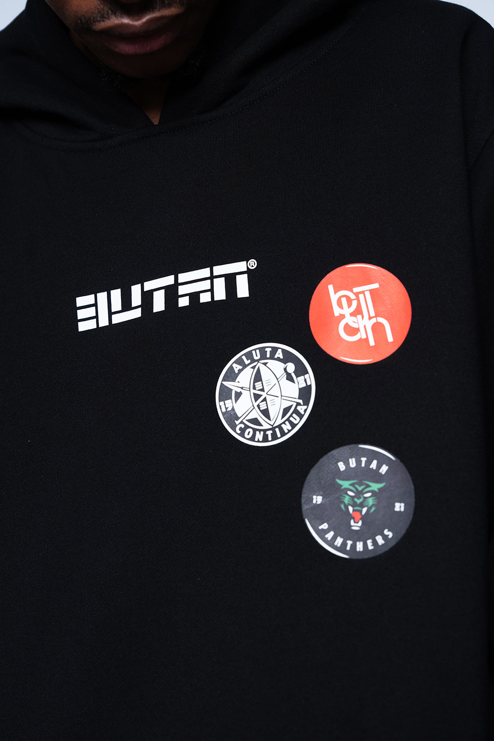 Close-up of a black hoodie featuring the 'BUTAN' brand logo in white on the chest, alongside three circular graphic patches in orange, white, and dark gray.