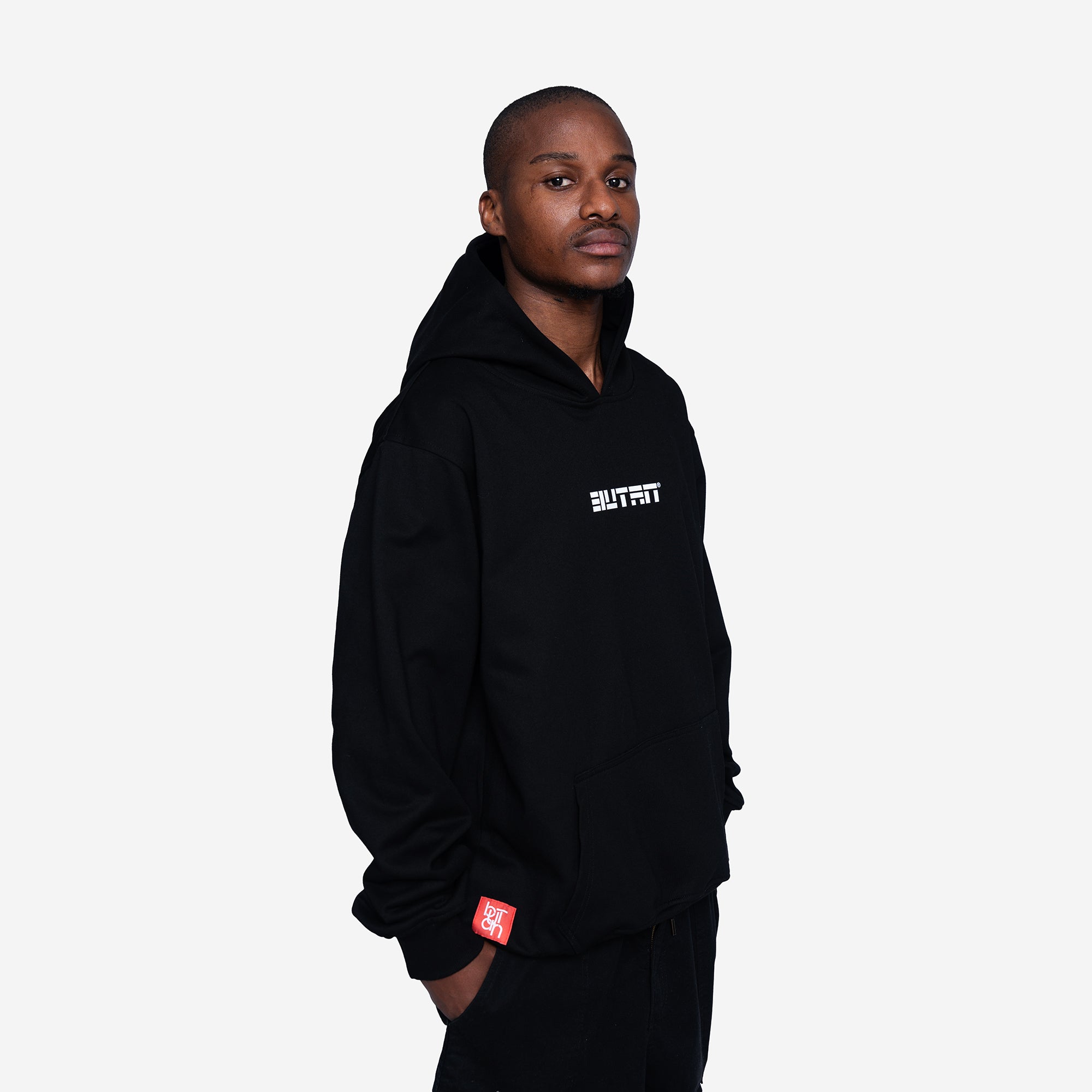 A man wearing a black pullover hoodie with a white 'BUTAN' logo on the chest and a small red tag near the cuff, photographed against a light gray studio background.