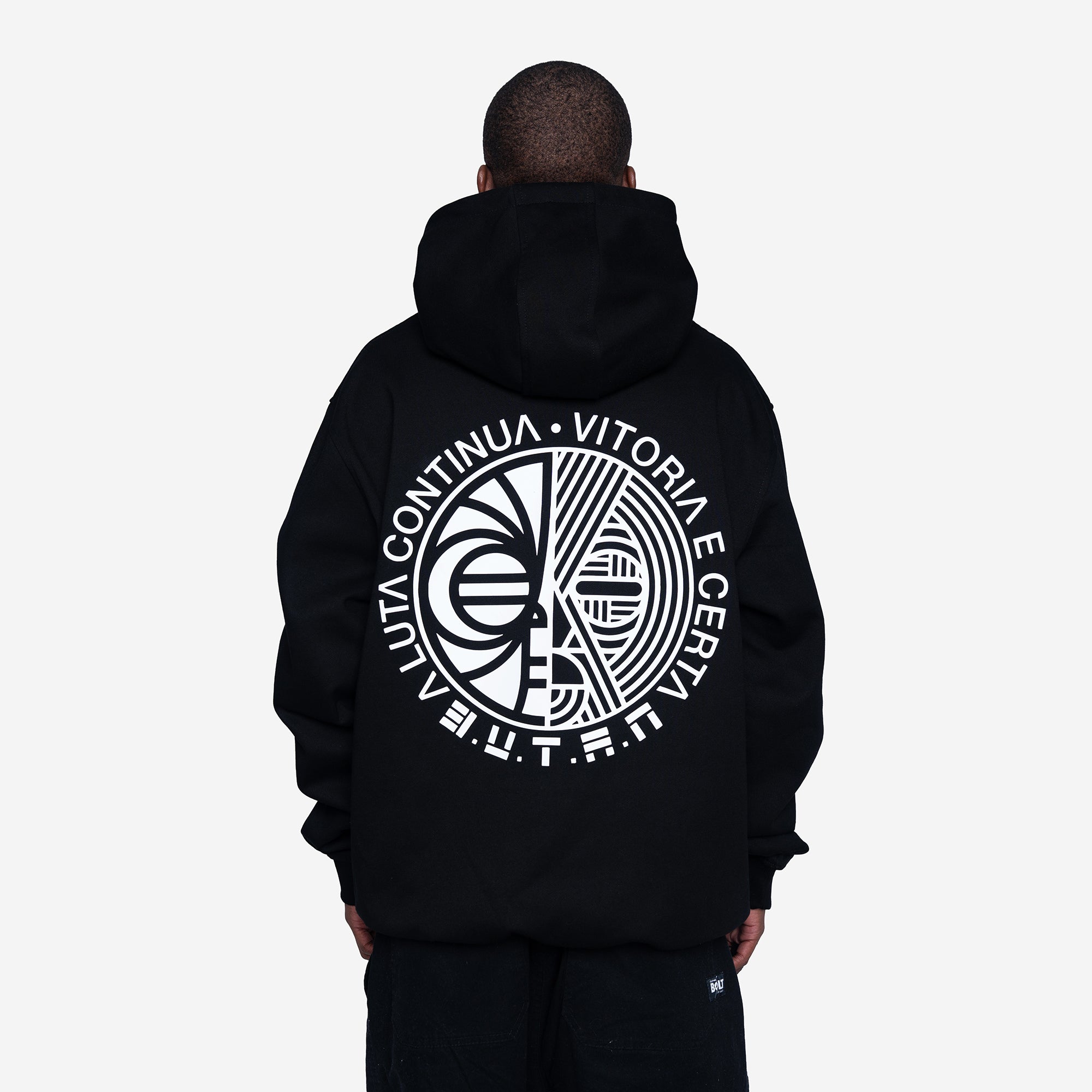 A person viewed from the back wearing a black pullover hoodie with a large white circular graphic and text printed across the back, against a light gray studio background.